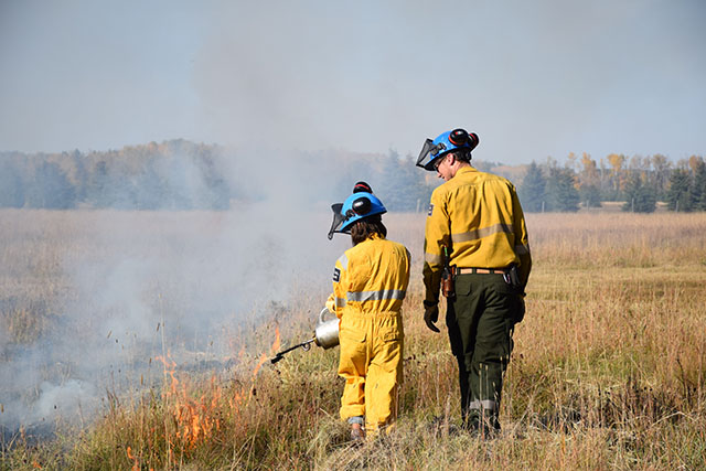 A youth and adult, both in protective jackets and helmets, walk side by side across a meadow. The youth is supervised igniting the grass.