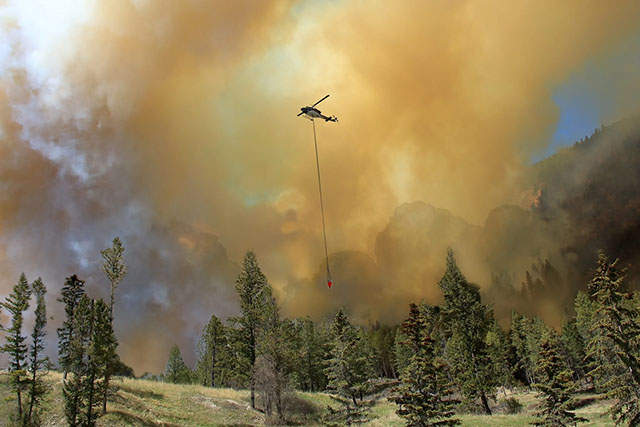 A prescribed fire burns in a forest, as a helicopter flies above the smoke with a suspended bucket of water.