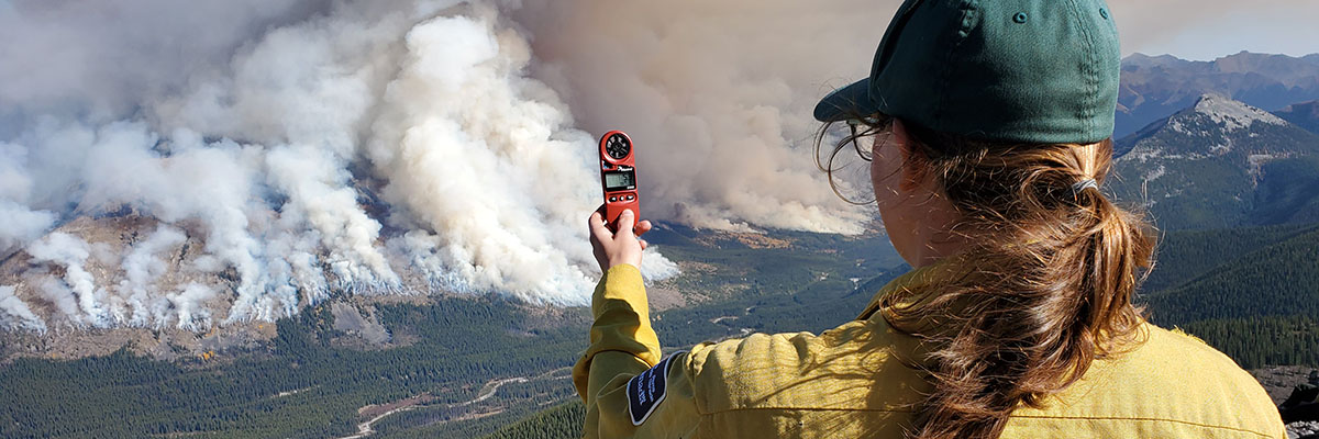 Kristyn Flanagan looks across a valley at a distant mountain range obscured by smoke and holds up a device to monitor weather conditions.