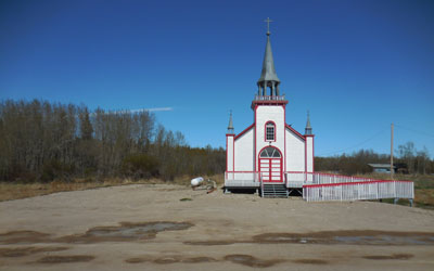 Vue générale de l’église anglicane St. Peter’s, qui montre sa masse rectangulaire coiffée d’un toit en pente et à pignon, et son porche de forme similaire, mais plus petit, surmonté d’un clocher. © Hay River Dene Band / Bande des Dénés de Hay River.