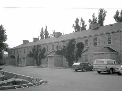 Vue générale de l'Édifice De Noyan, montrant son échelle qui est celle d’une habitation et la simplicité de la conception, soit une structure rectangulaire de deux étages coiffée d’un toit à pignon à pan coupé qui est percé par des cheminées en pierre, 19 (© (Environment Canada, Canadian Parks Service, Architectural History Branch/ Environnement Canada, Service canadien des parcs,  Direction de l'histoire de l'architecture, J. Adell, 1989.))