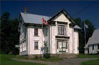 Vue générale de la mairie de Bolton-Est, qui montre sa construction à ossature de bois, revêtue de bardage à clin peint en blanc. (© Parks Canada Agency / Agence Parcs Canada.)