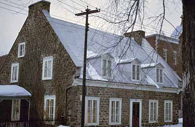 Corner view of Hertel de la Fresnière House at Trois-Rivières Historical Complex, showing the gable roof with dormer windows and twin end-chimneys. © Parks Canada Agency / Agence Parcs Canada