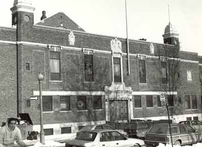 Vue de l'entrée principal du manège militaire, qui montre le caractère de style néoclassique édouardien de la façade, soit l’entrée principale formée par un grand portail en bois et les deux petites tours à dôme en cuivre, 1993. © Department of National Defence / Ministère de la Défense nationale, 1993.